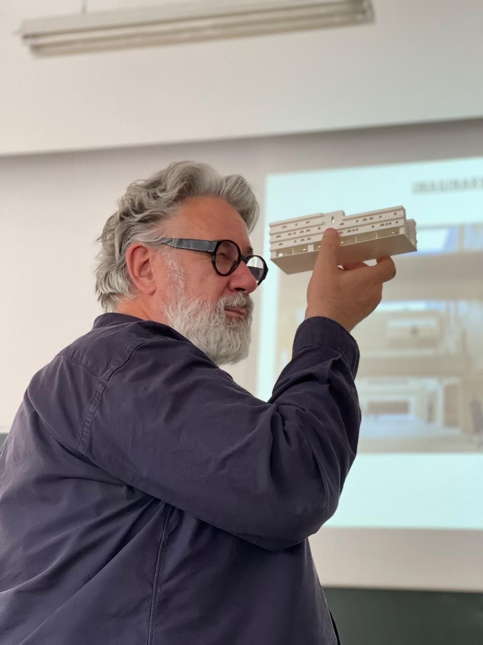 Man holding up a model of a structure in front of a projection screen in a classroom setting.