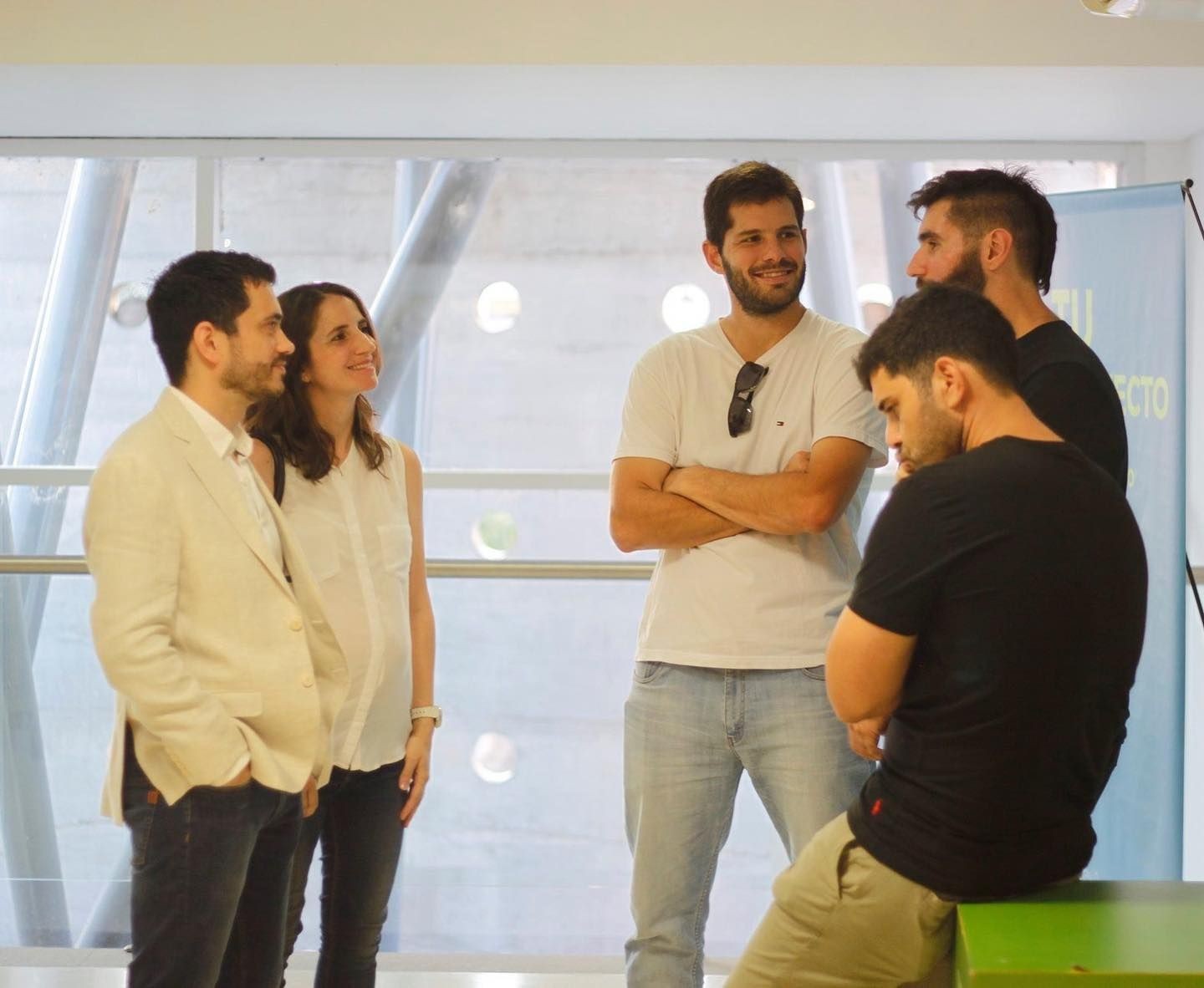 Group of five people engaged in conversation, standing near large windows in a brightly lit room.